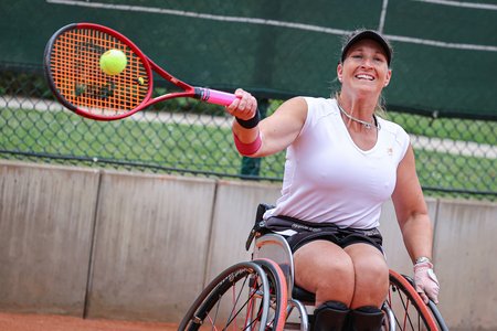 Eine Frau sitzt im Rollstuhl und spielt auf Schulterhöhe einen Tennisball mit einem Schläger.