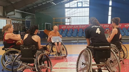 Vier Rollstuhlnutzerinnen und ein Trainer - ebenfalls im Rolli - sitzen im Kreis in einer Sporthalle. Der Coach hat eine Taktiktafel in der Hand und erklärt.