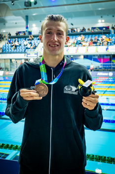 Ein junger Mann mit schwarzer Trainingsjacke zeigt seine Bronzemedaille in die Kamera. Im Hintergrund befindet sich ein Schwimmbecken.
