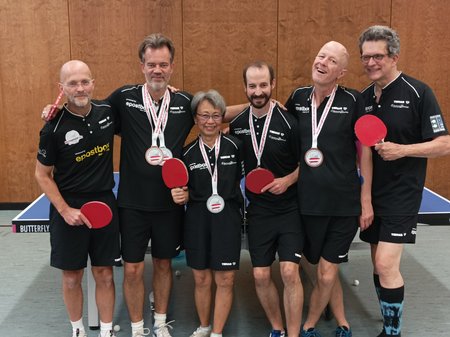 Sechs Personen in schwarzer Sportkleidung posieren mit Medaillen und Tischtennis-Schägern für ein Foto.