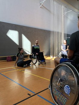 Eine Frau sitzt in einer Sporthalle auf dem Boden, daneben ein Mann im Rollstuhl. Im rechten Bildanschnitt ist ein Reifen eines Rollstuhls zu sehen.