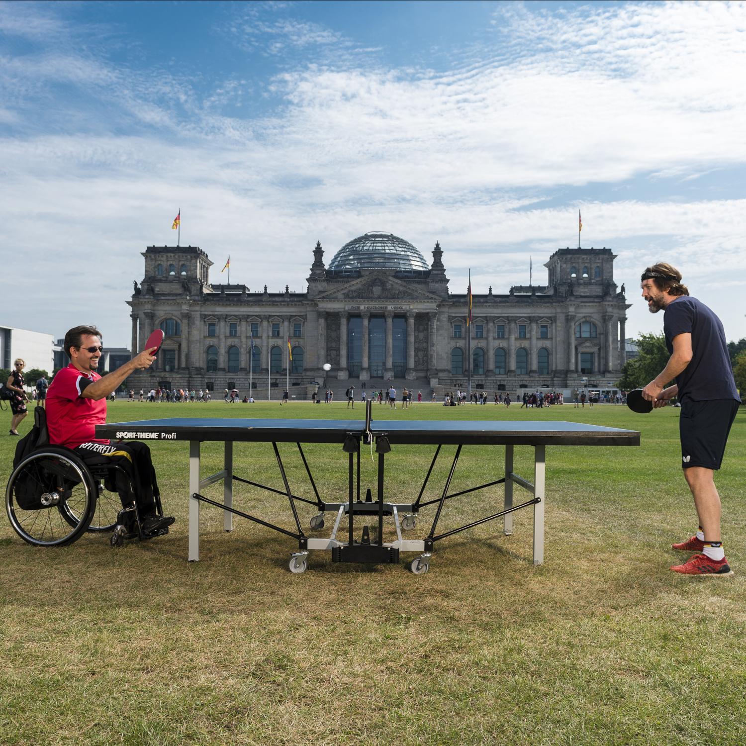 Rollstuhlfahrer spielt mit Fußgänger Tischtennis, im Hintergrund mittig der Reichstag.