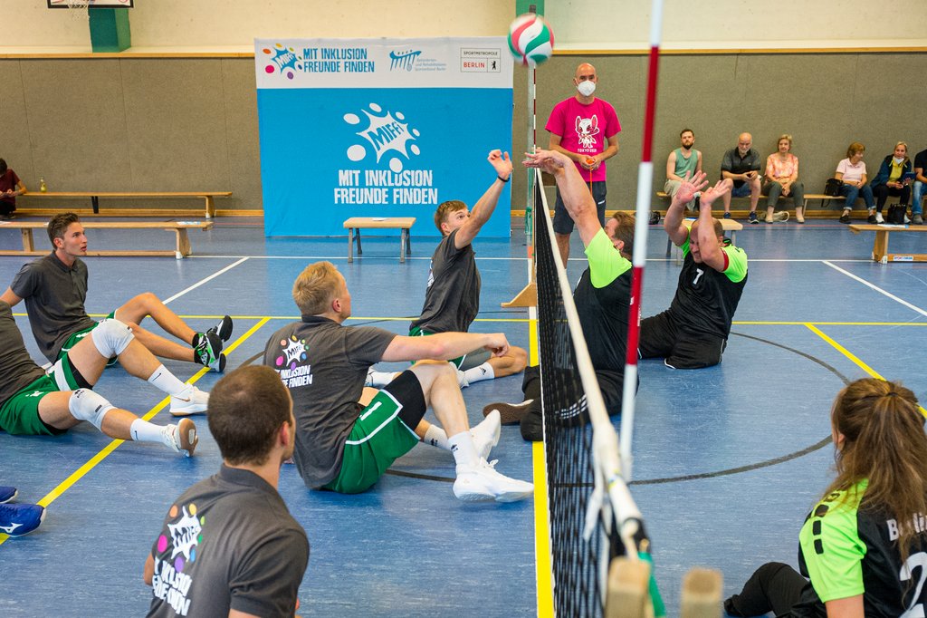 Menschen sitzen am Hallenboden und haben die Hände nach oben gerichtet. In der Mitte zwischen den Mannschaften ist ein Netz. Ein Volleyball befindet sich über dem Netz.