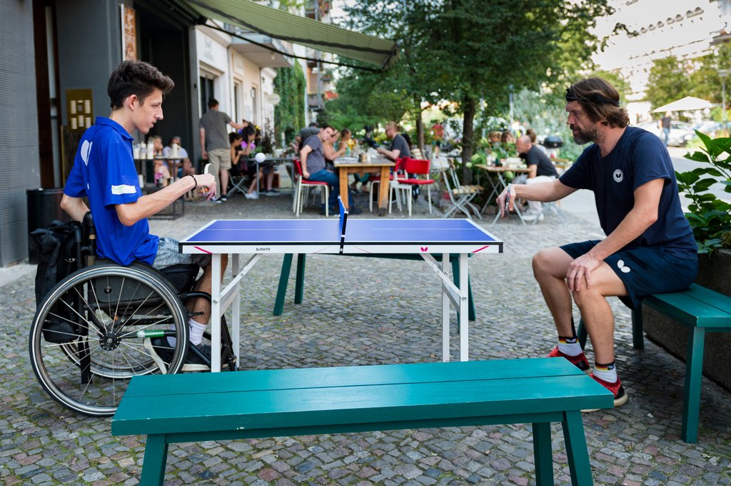Zwei junge Männer spielen auf einer verkleinerten Tischtennisplatte. Der linke Mann sitzt im Rollstuhl.