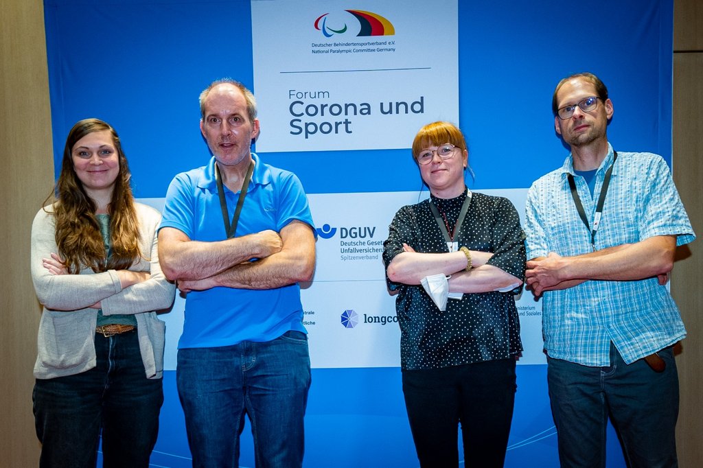 Die vier genannten Personen stehen lächelnd mit verschränkten Armen vor einer Werbewand mit der Aufschrift "Corona und Sport".